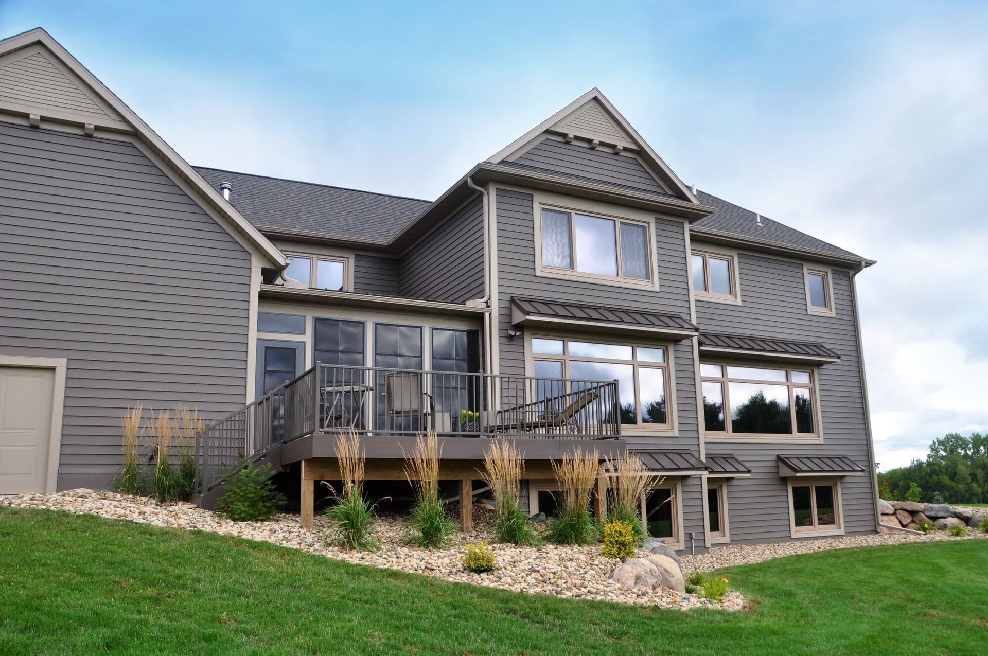 Gray two-story house with a deck, windows, and landscaping, under a cloudy sky.
