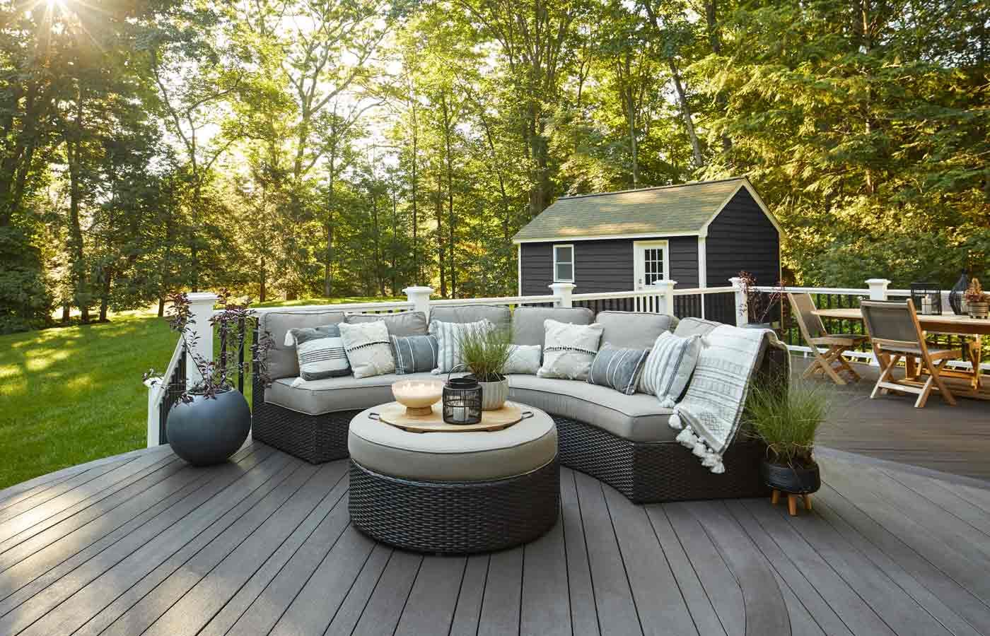 Outdoor deck with gray sectional sofa, ottoman, and small black shed. Green trees in background.