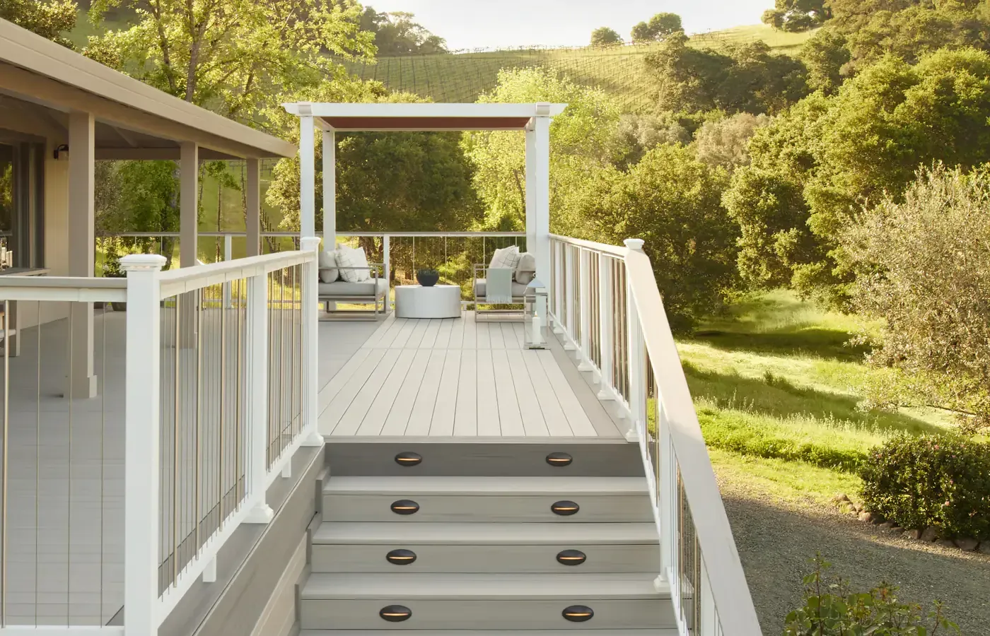 A deck with white railing and steps leads to seating area, overlooking a green landscape.