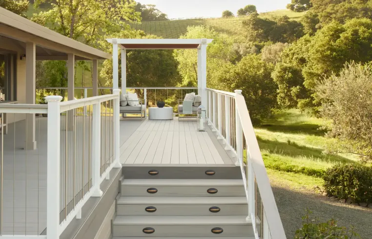 A deck with white railing and steps leads to seating area, overlooking a green landscape.