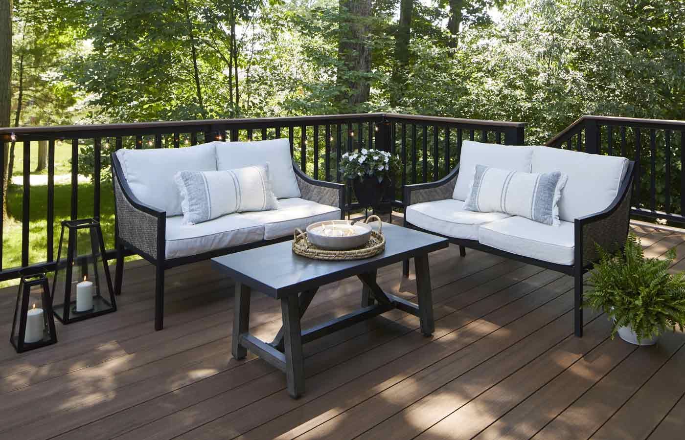 Outdoor deck with seating: two loveseats with white cushions and pillows, gray coffee table, black lanterns, surrounded by trees.