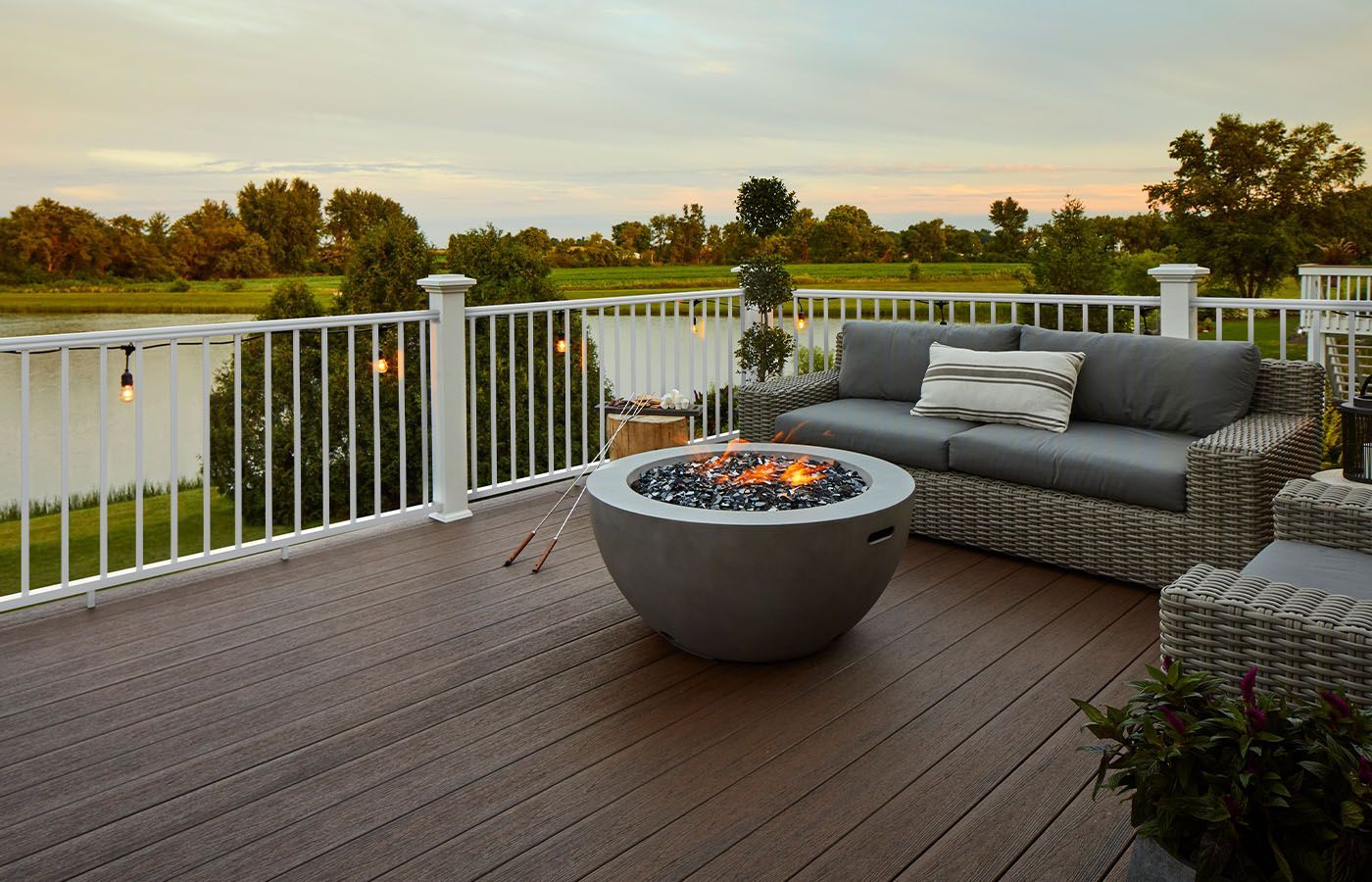 Deck with fire pit, gray wicker furniture, and white railing overlooking a lake and trees at sunset.