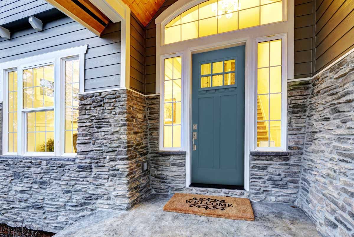 Blue front door with sidelights and arched transom, gray stone facade and welcome mat.