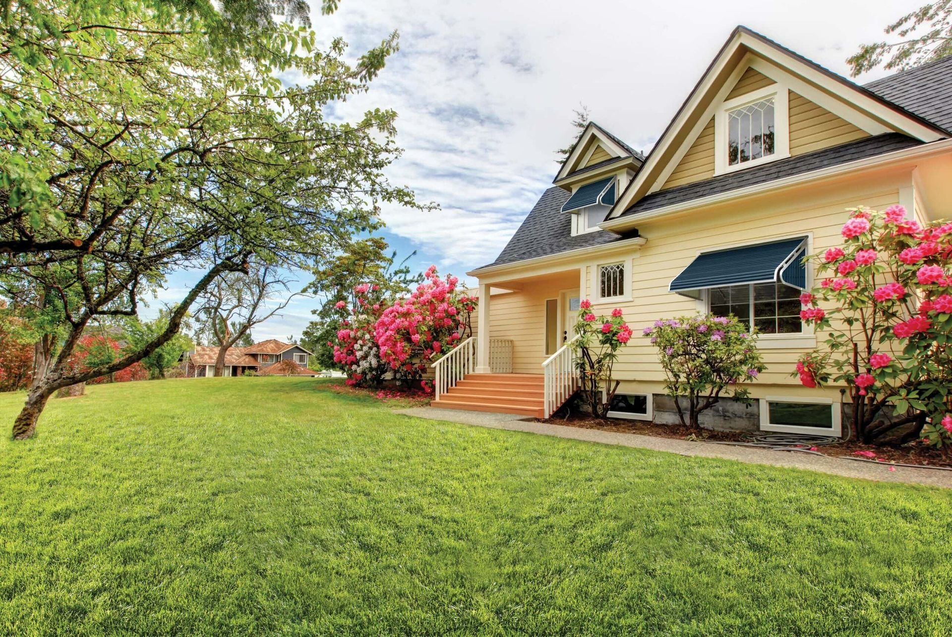 Yellow Victorian house with green lawn, pink roses, and cloudy sky.