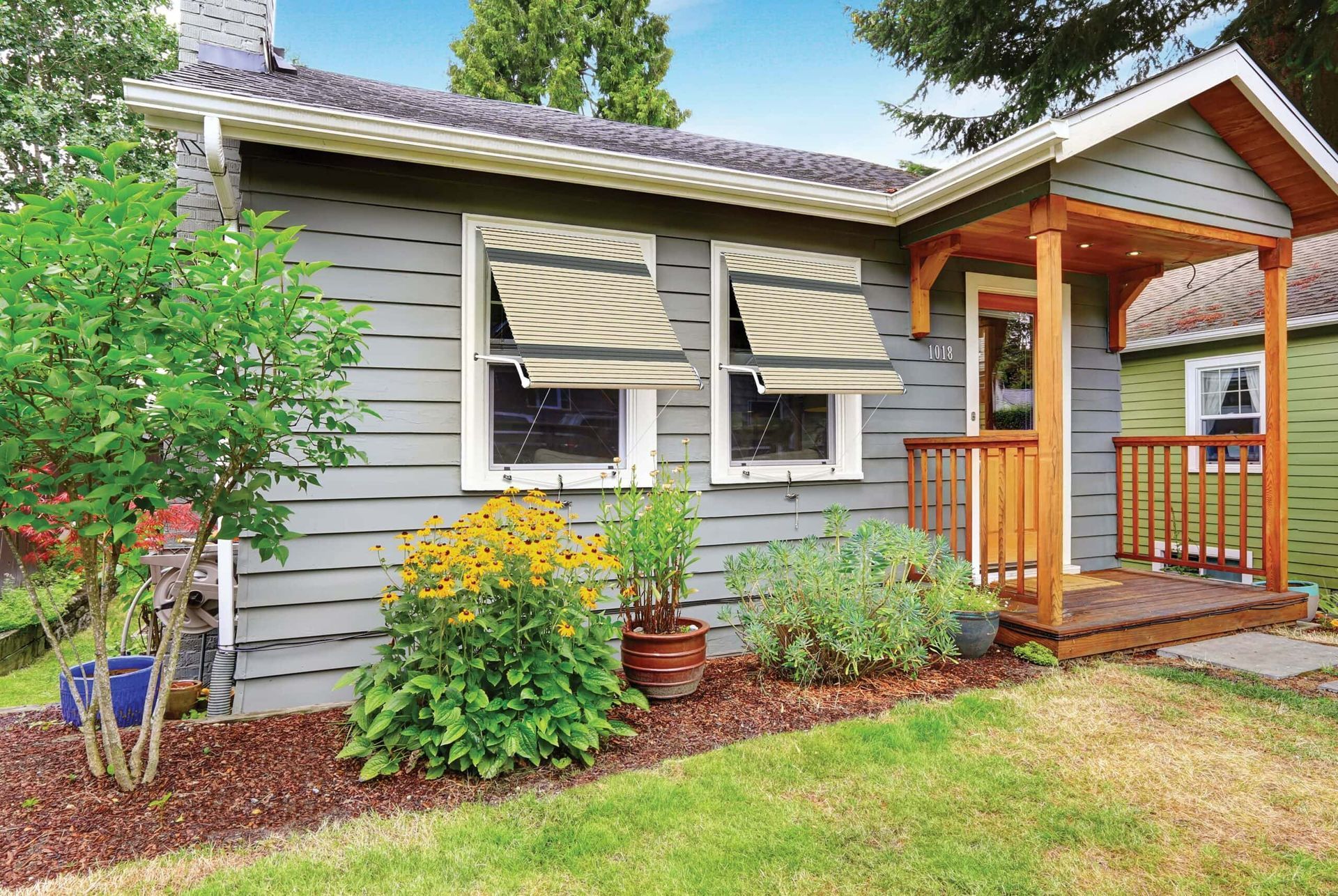 A small gray house with tan window awnings, a porch, and a garden.