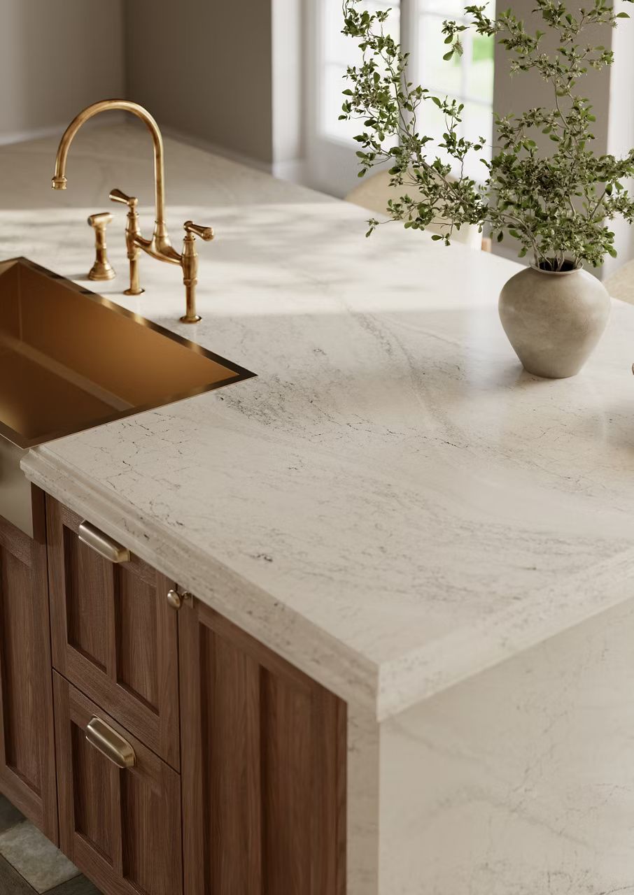 Kitchen island with light countertops, bronze sink and faucet, and wooden cabinets. Vase with greenery.