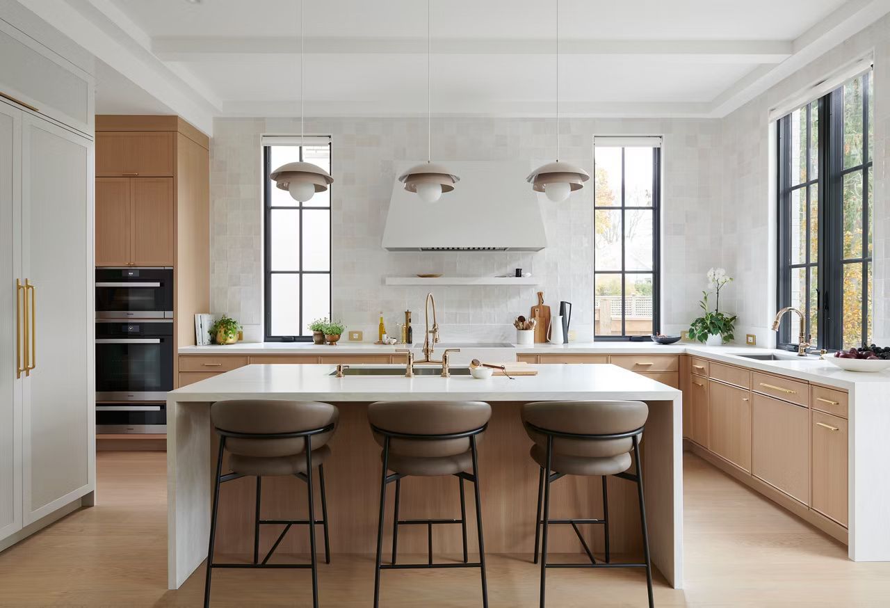 Modern kitchen with wood cabinets, white countertops, island with stools, and large windows.