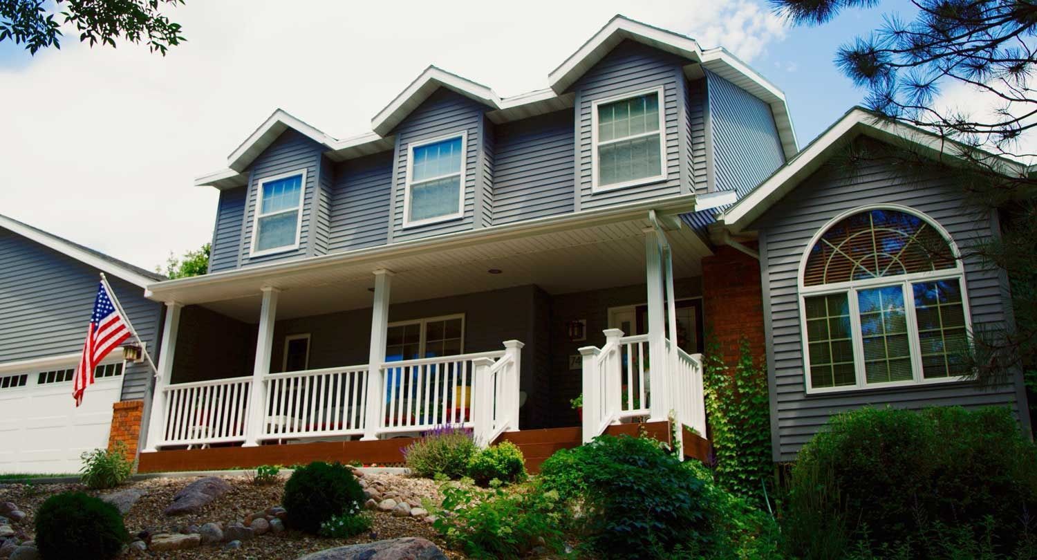Two-story gray house with white porch and three dormers, American flag, sunny day.