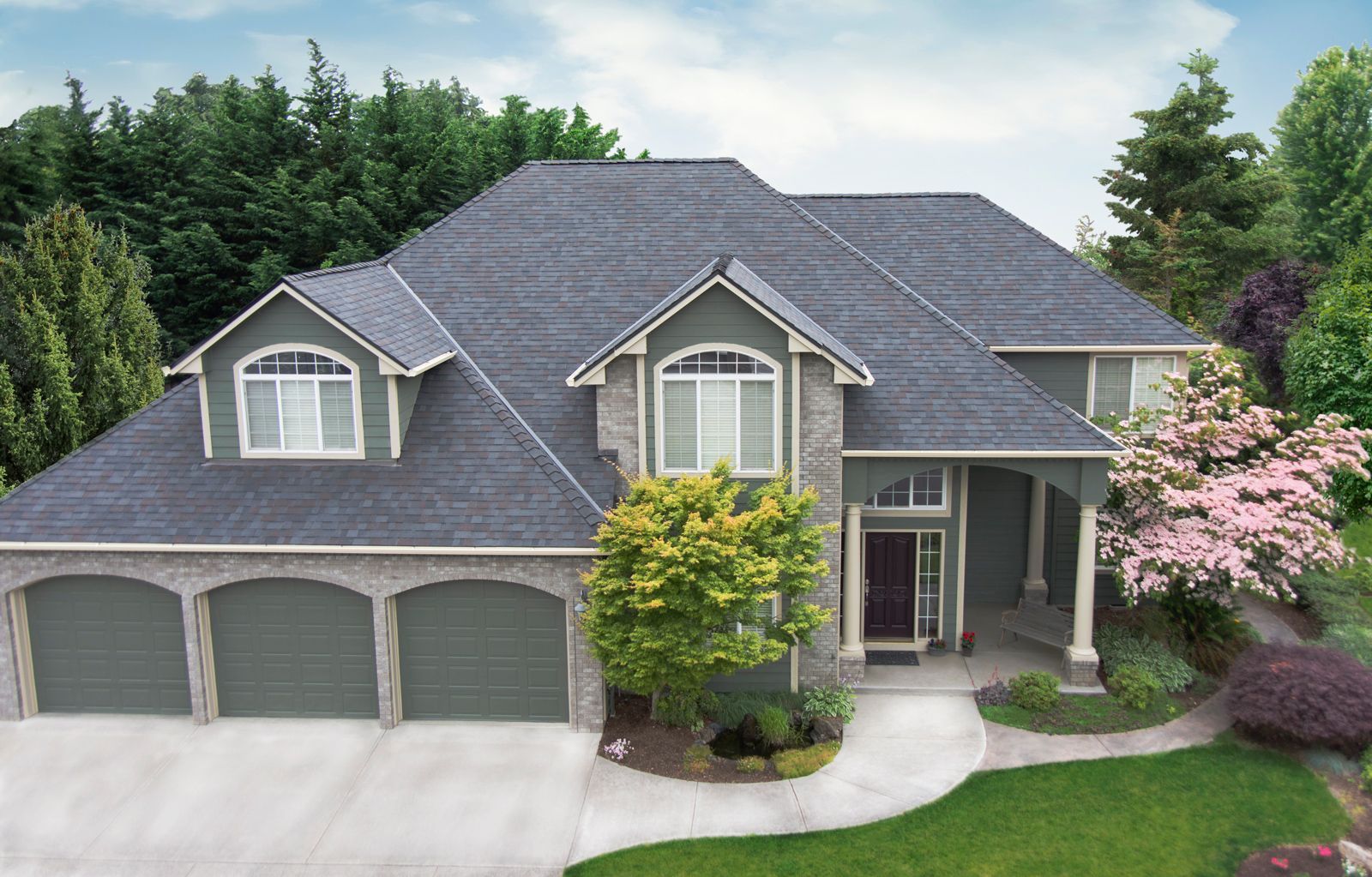 Two-story house with green siding, gray roof, stone accents, three-car garage, surrounded by greenery.