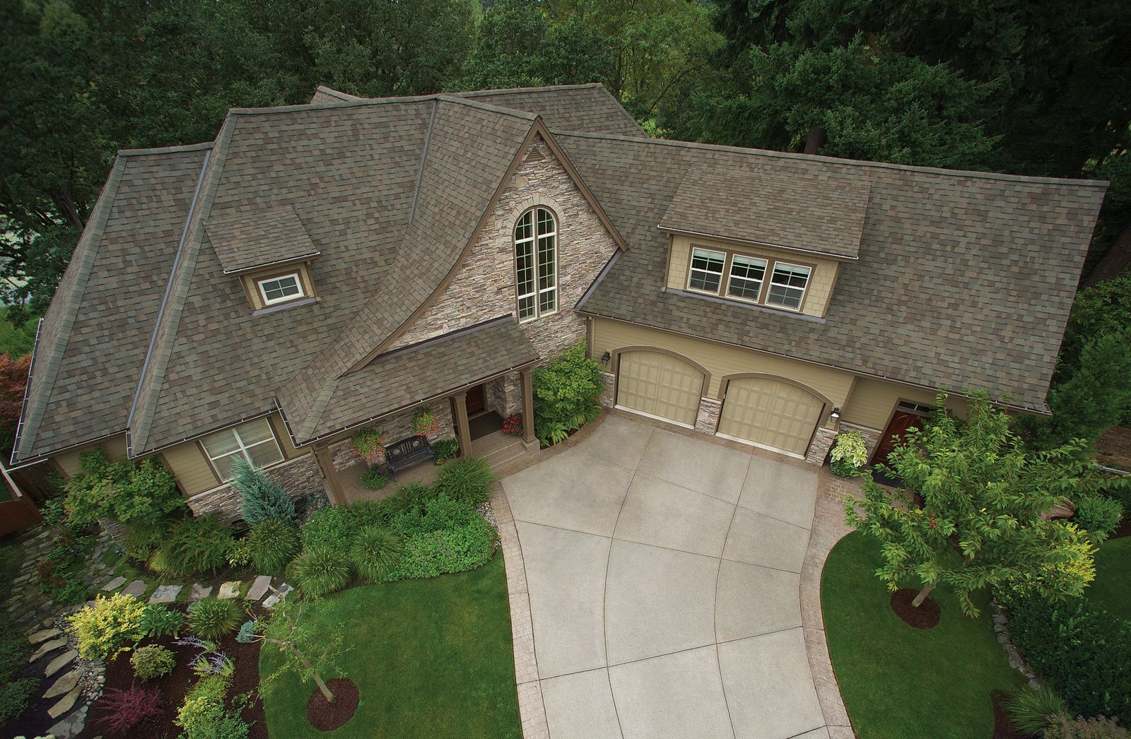 Aerial view of a large, tan house with stone accents, a driveway, and lush landscaping surrounded by trees.