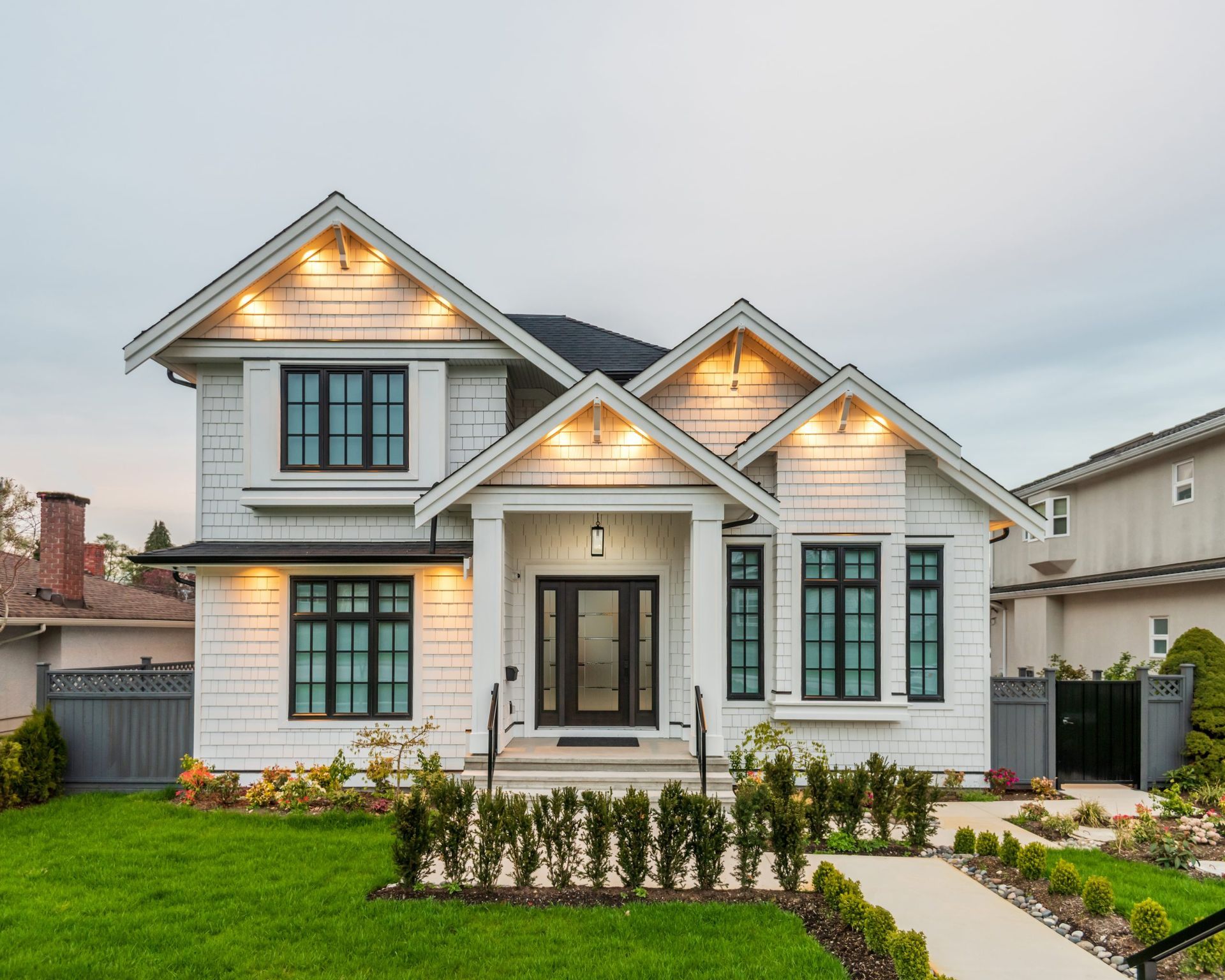 White two-story house with black trim, lit with warm lights, set on a green lawn with a pathway.