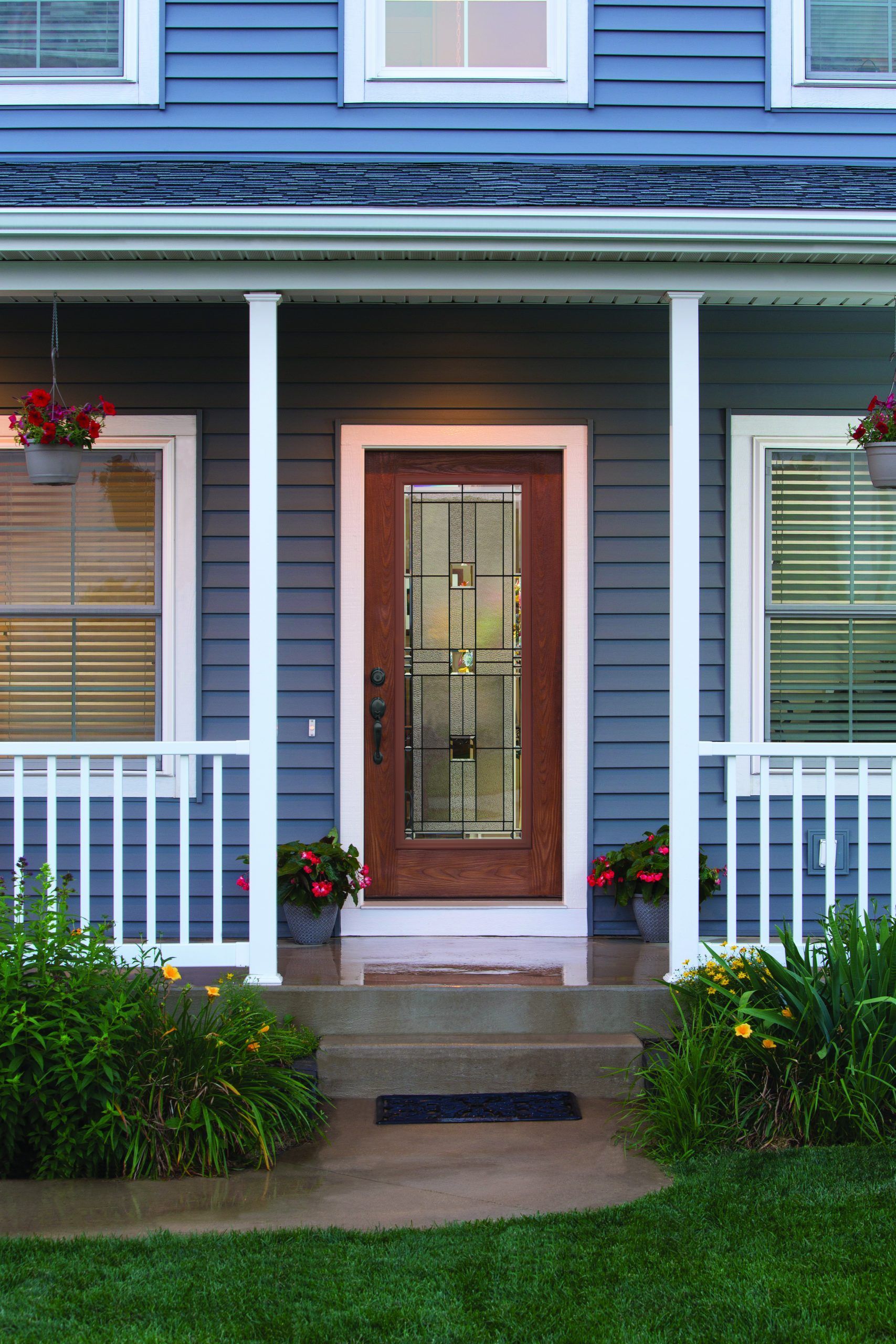 Blue house with white porch, brown door, and plants; flower pots hang on the porch.
