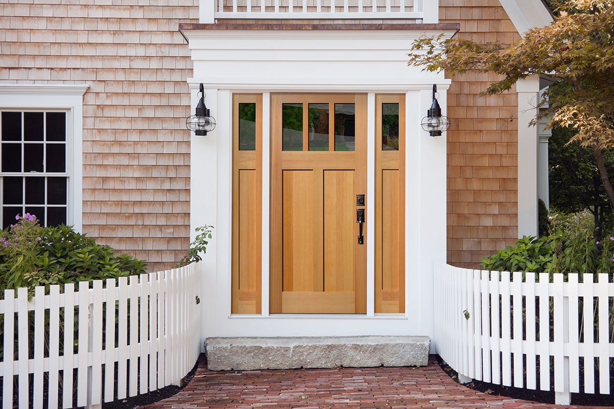 Wooden door with sidelights in a white frame, between a white picket fence, in a shingled house.