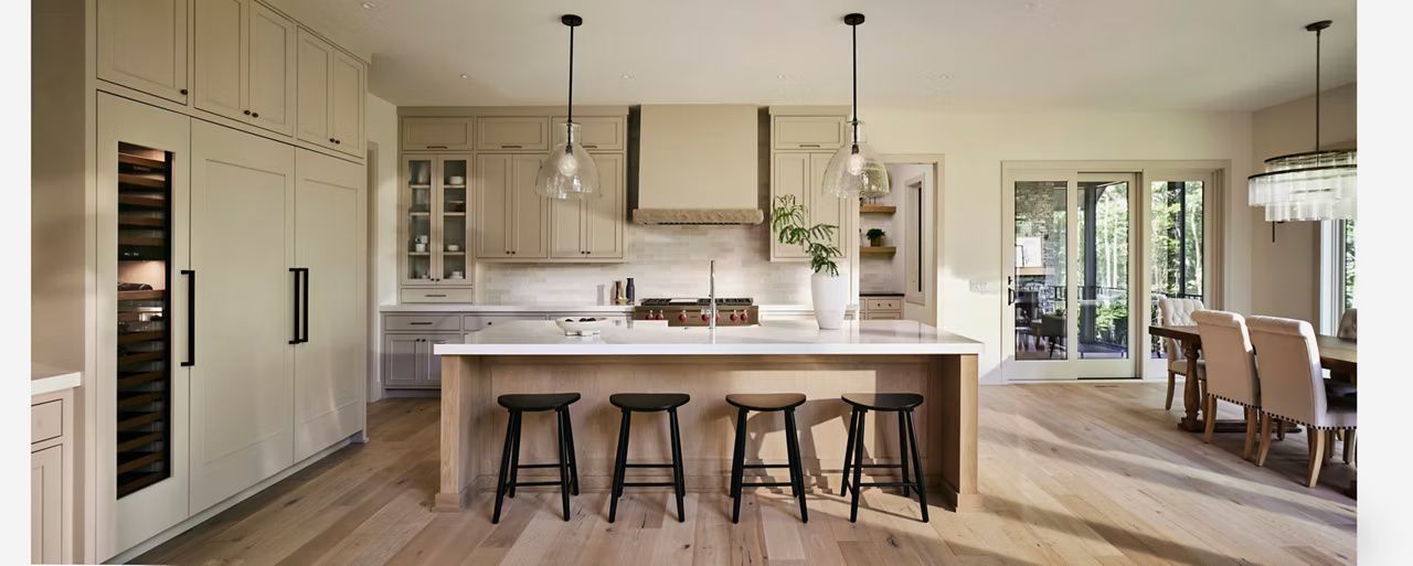 A modern kitchen with a large island and dining area. Light wood cabinets, black stools, and pendant lights.