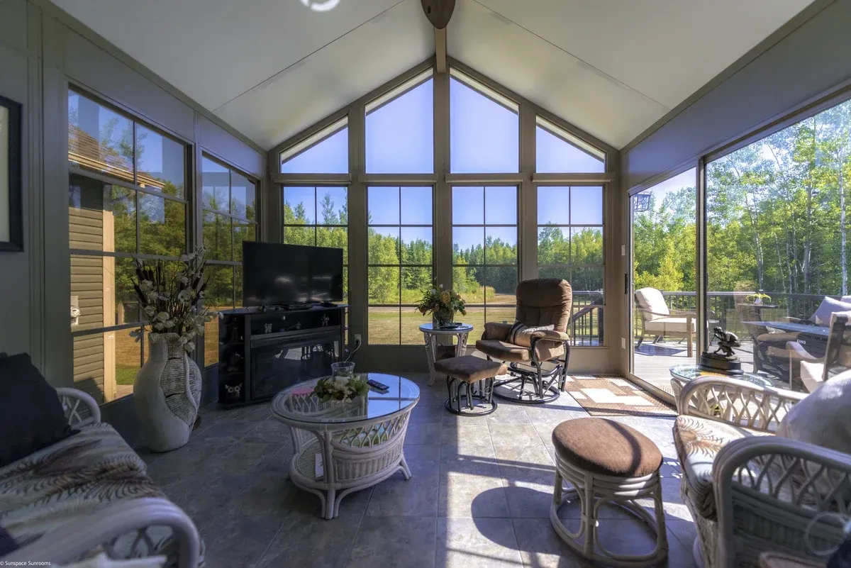 Sunroom with wicker furniture, TV, and large windows overlooking trees.