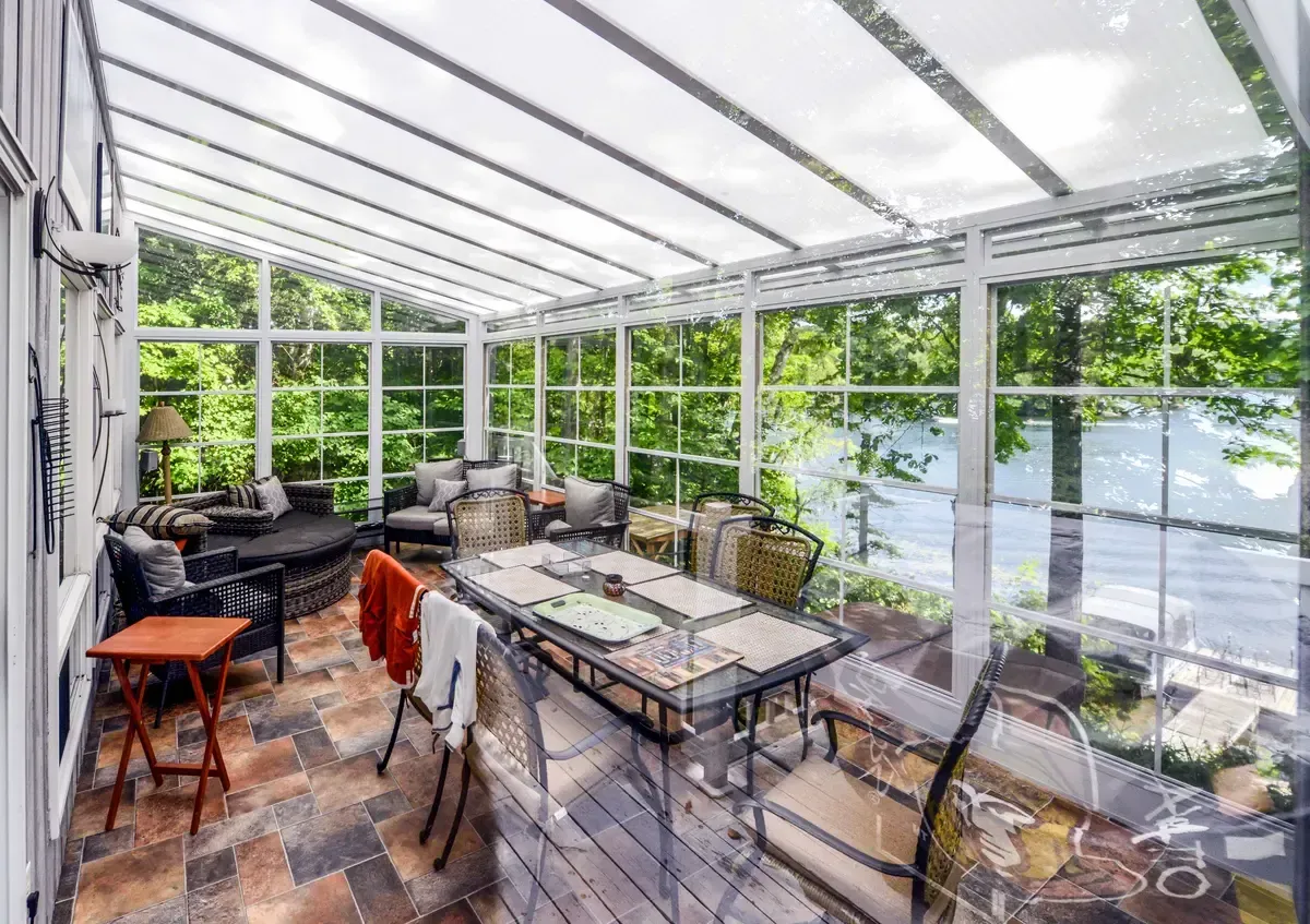 Sunroom with glass walls, a table, chairs, and outdoor view. Sunlight streams in.