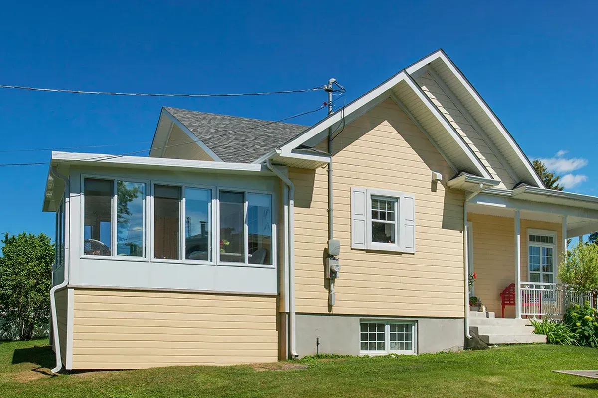 Yellow house with sunroom and porch on a green lawn under a blue sky.