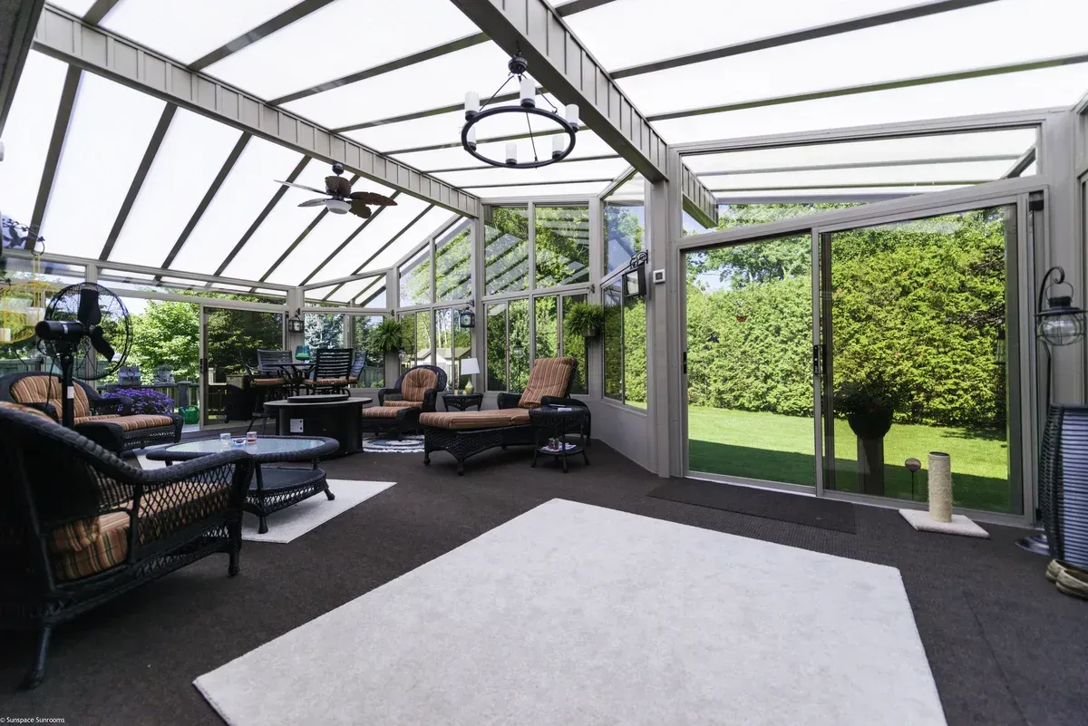 Sunroom with glass walls and roof, furnished with wicker furniture, overlooking a backyard.