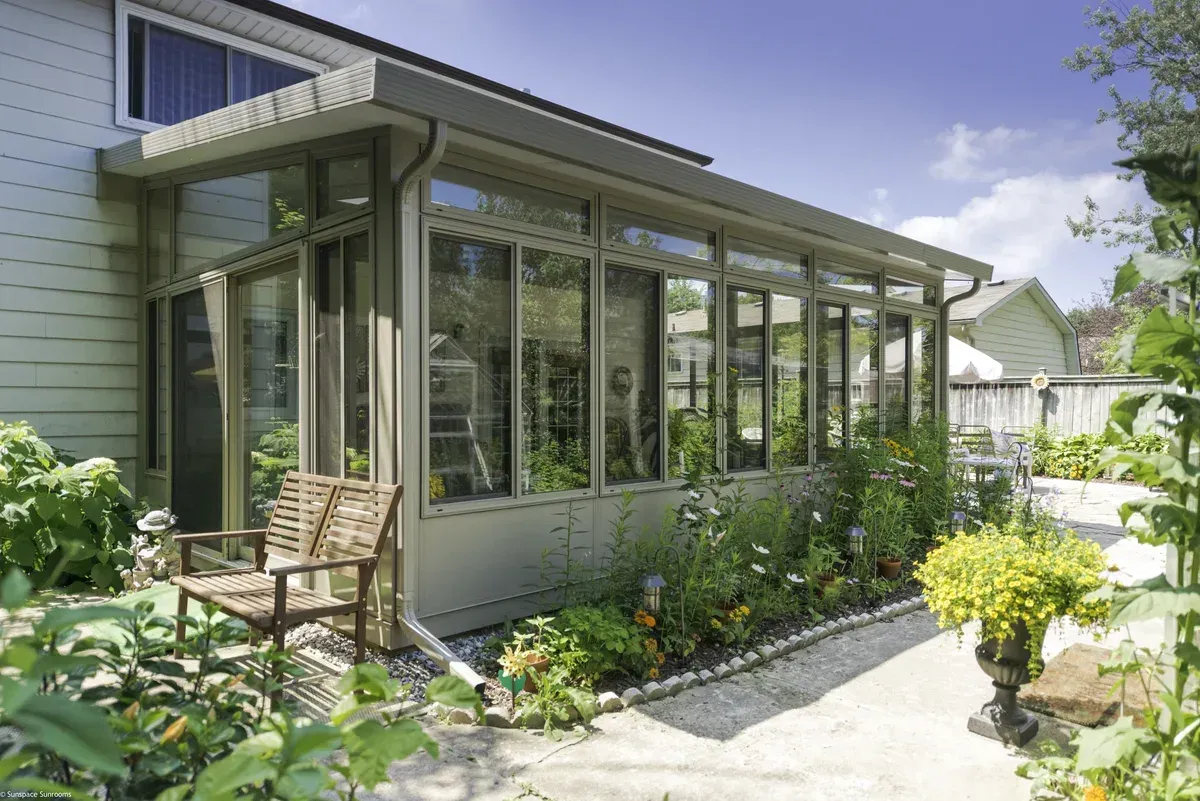 Sunroom addition attached to a house, with glass walls, surrounded by a garden and plants.