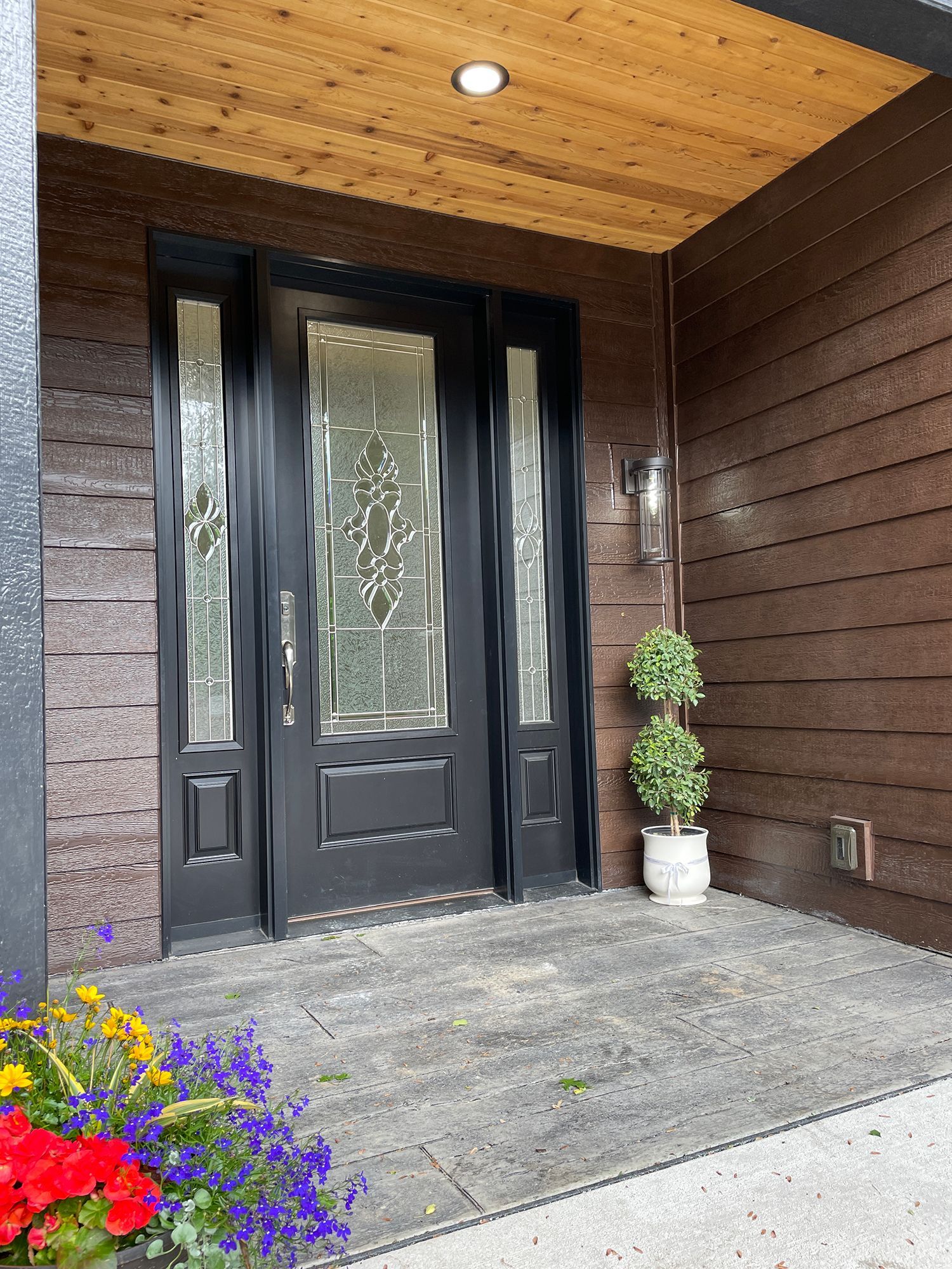 Black front door with glass panels, wood siding, and a small topiary on a porch.