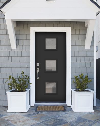 Black door with three square windows, white trim, gray siding, white planters with greenery.