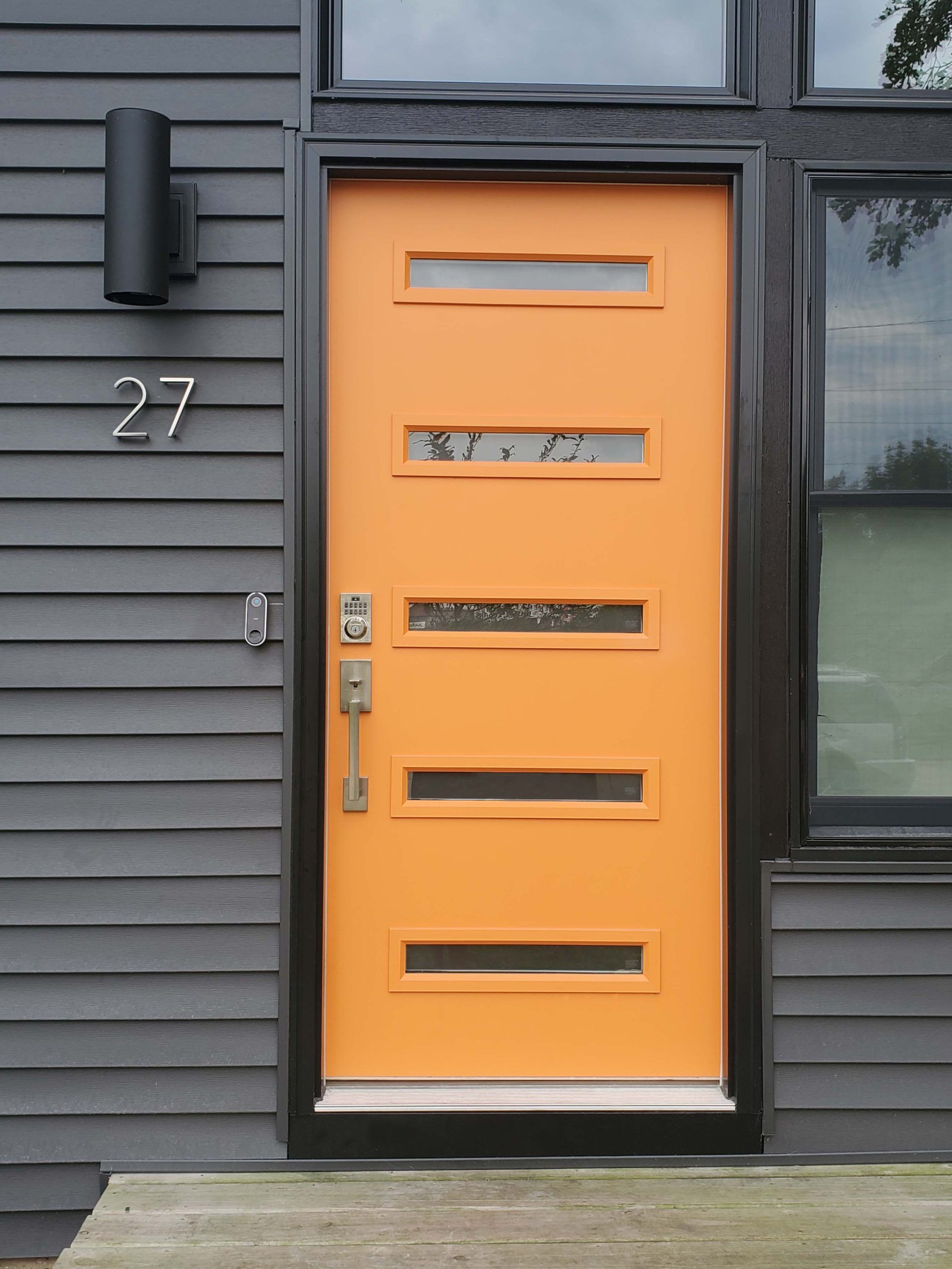 Bright orange door with black trim, horizontal windows, on a gray siding house; number 27.