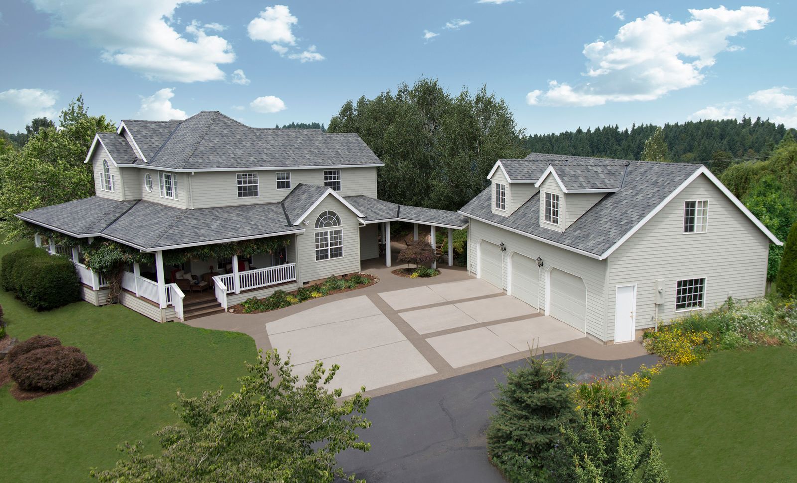Two-story light gray house with attached garage and large driveway, surrounded by green lawn and trees under blue sky.