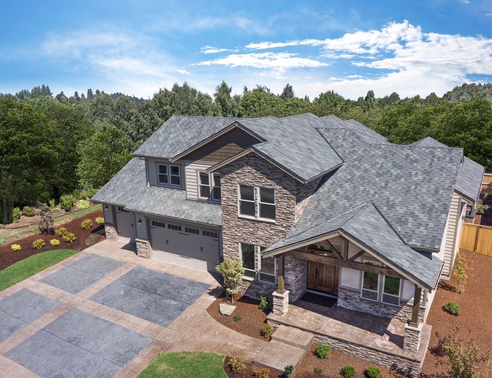 Two-story house with stone and gray siding, driveway, and a tree-lined background under a blue sky.