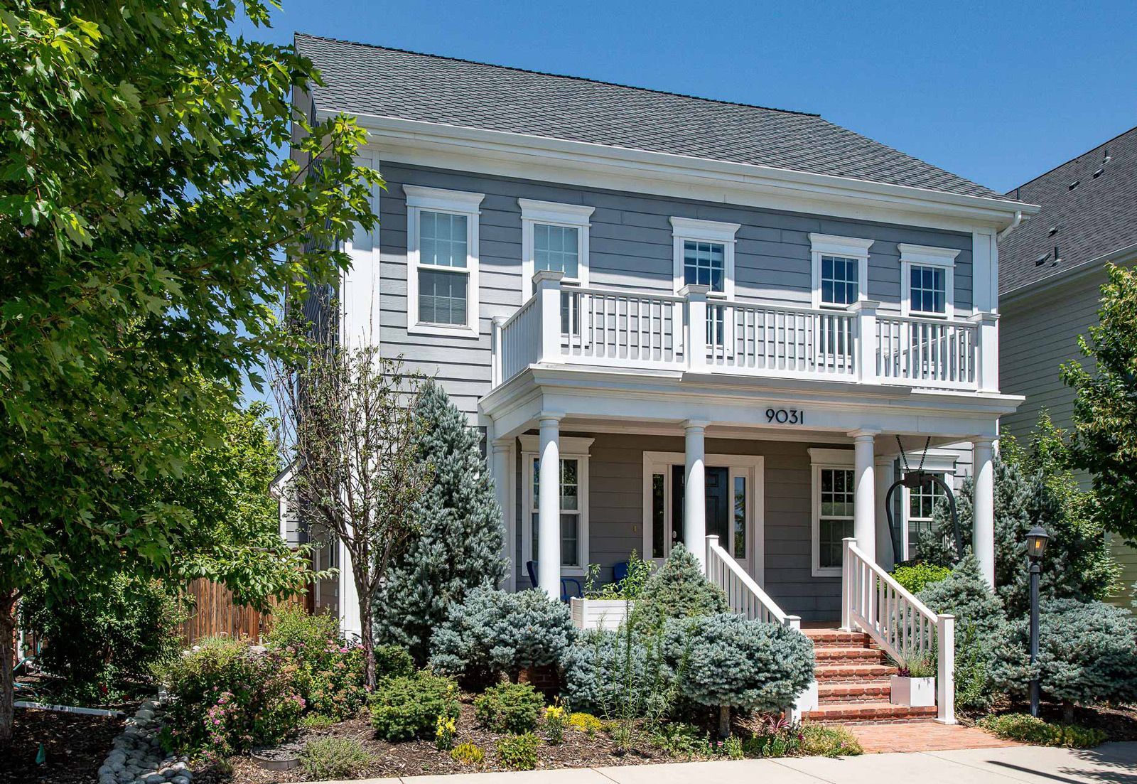 Two-story gray house with white trim and a balcony, surrounded by trees and bushes.
