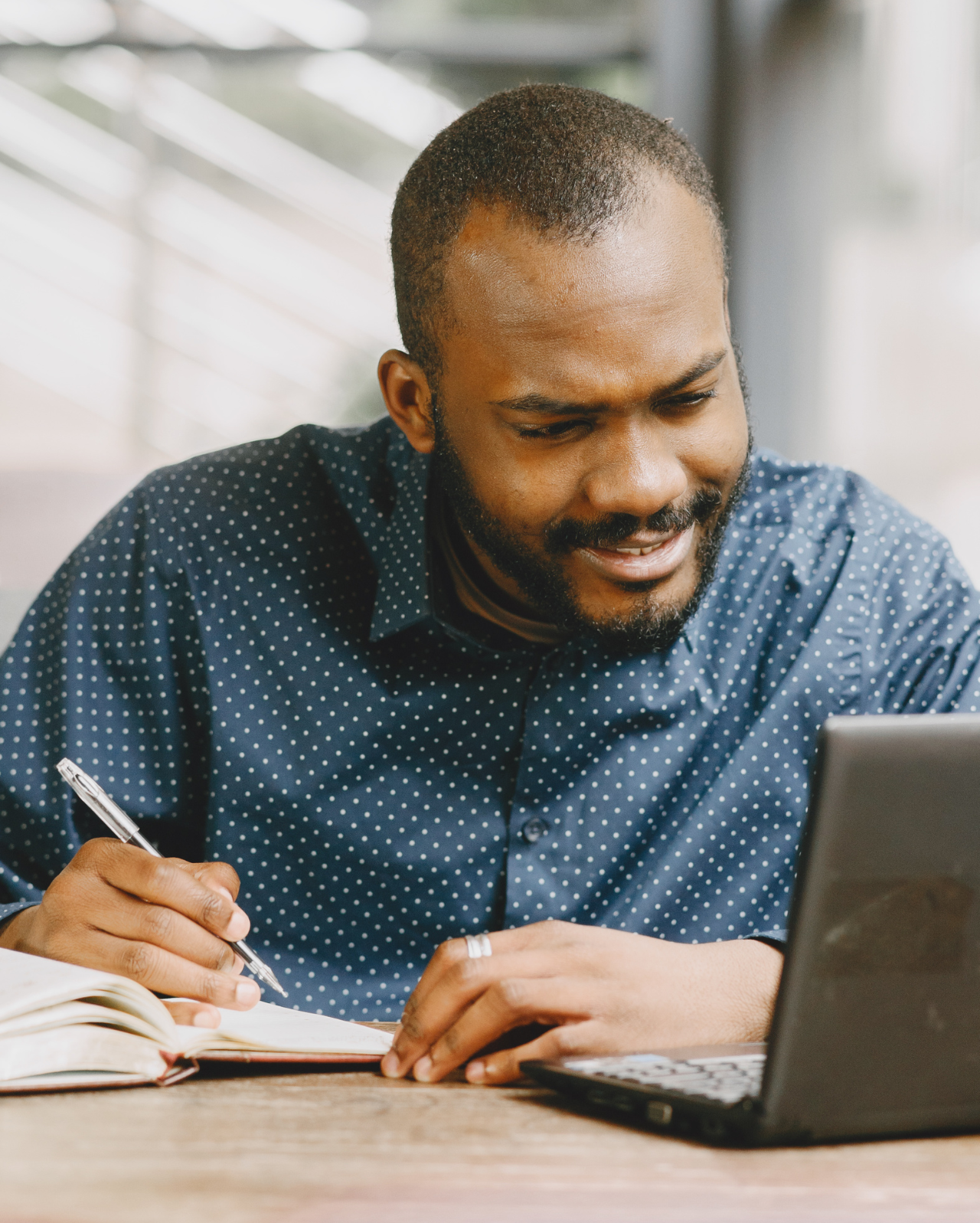 A man is writing in a notebook while using a laptop computer.