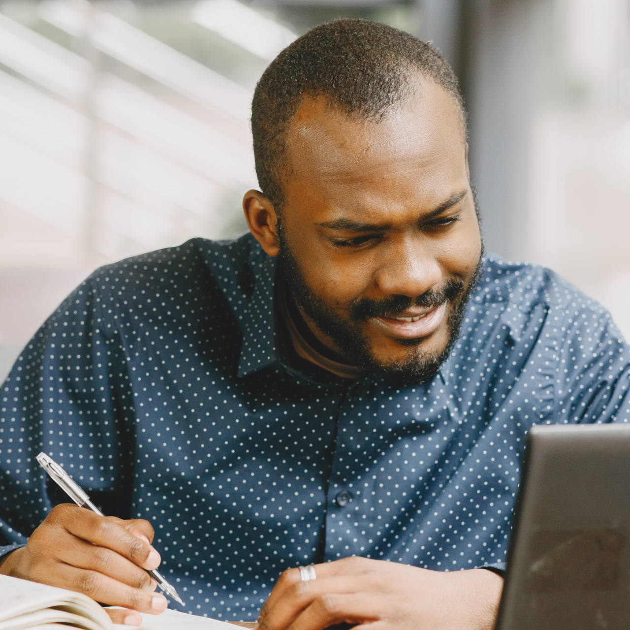 A man is writing in a notebook while looking at a laptop