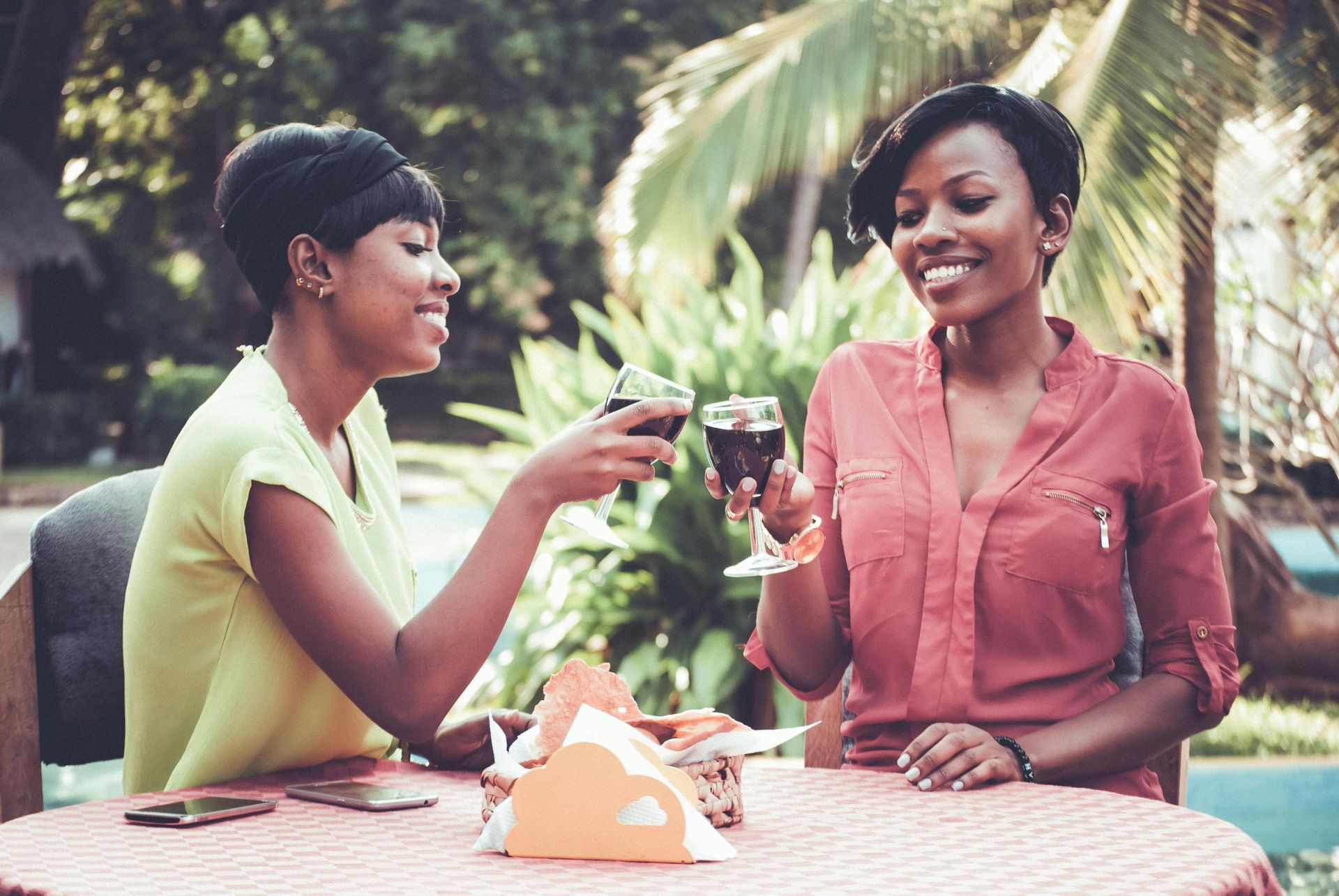 Two people sitting at an outdoor table, smiling and clinking glasses of red wine together.