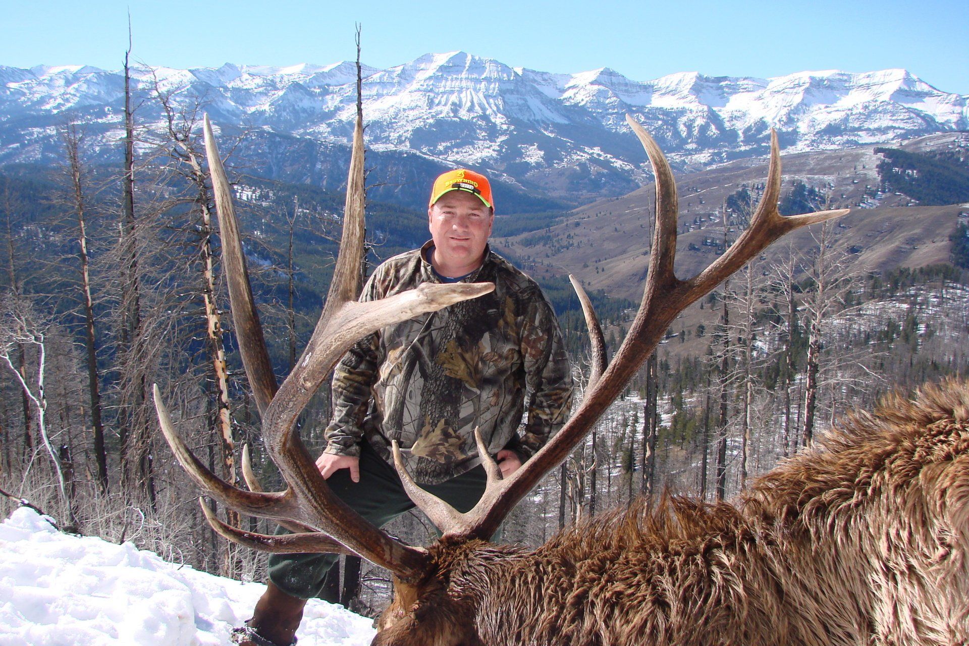 Man in camo with a large elk, antlers framing him, snowy mountain backdrop.