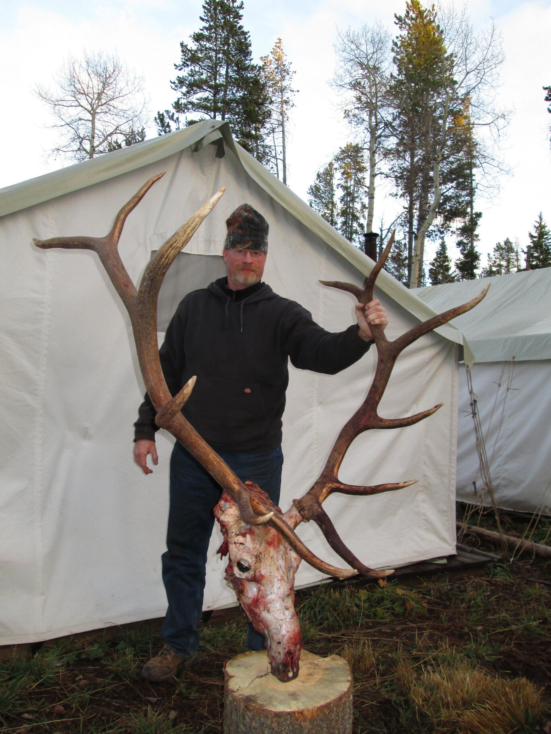 Man holding large elk antlers next to a tent. The skull is on a tree stump in a forest setting.