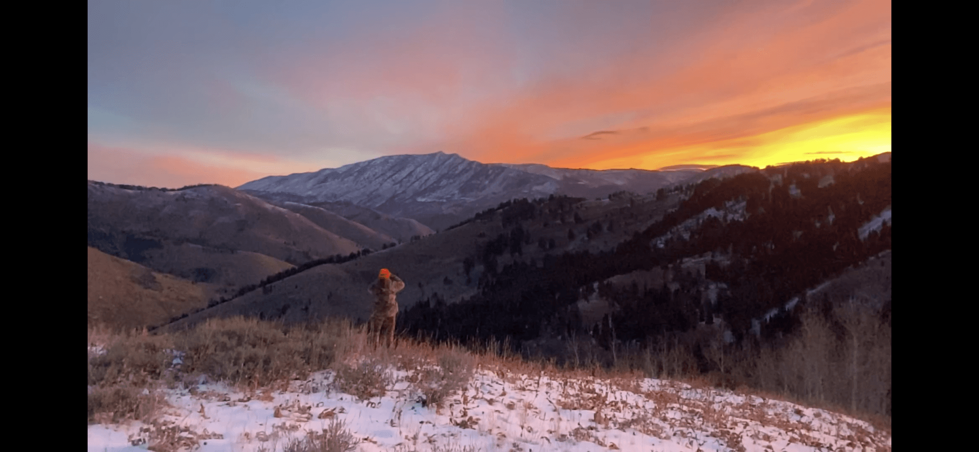 A person is standing on top of a snowy mountain at sunset.