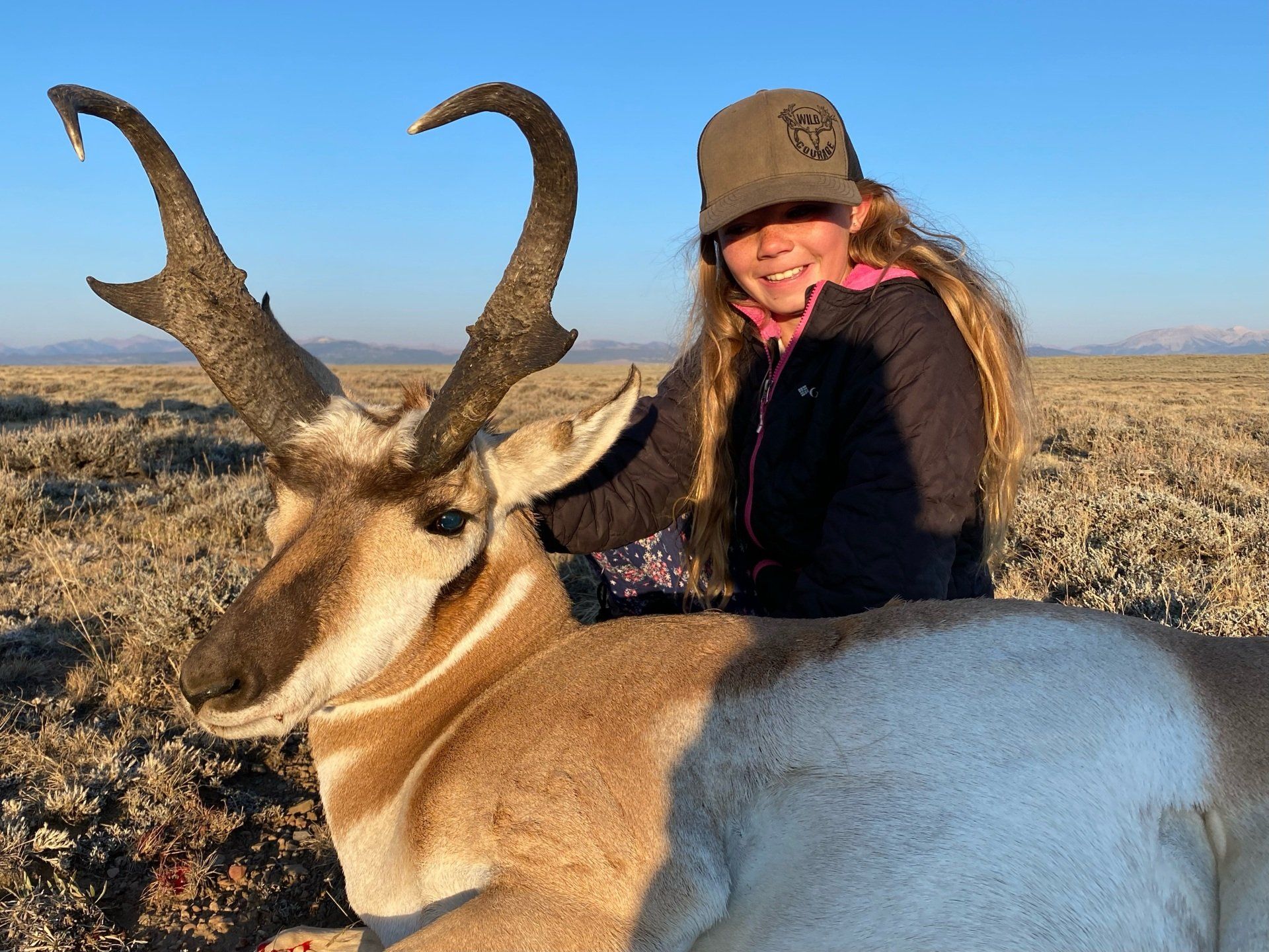 A woman in a hat stands next to a large antelope