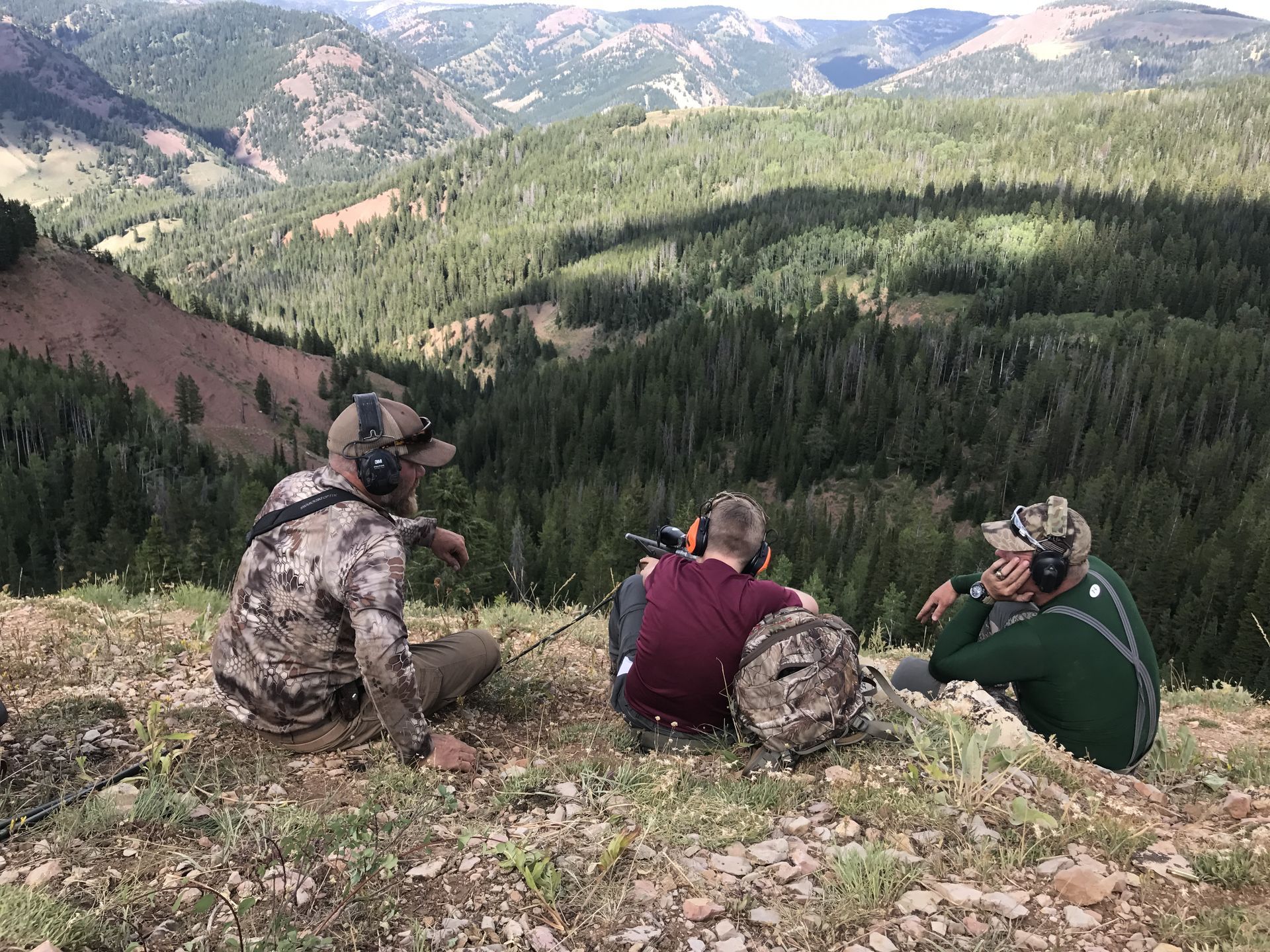 Three men are sitting on top of a mountain looking out over a forest.