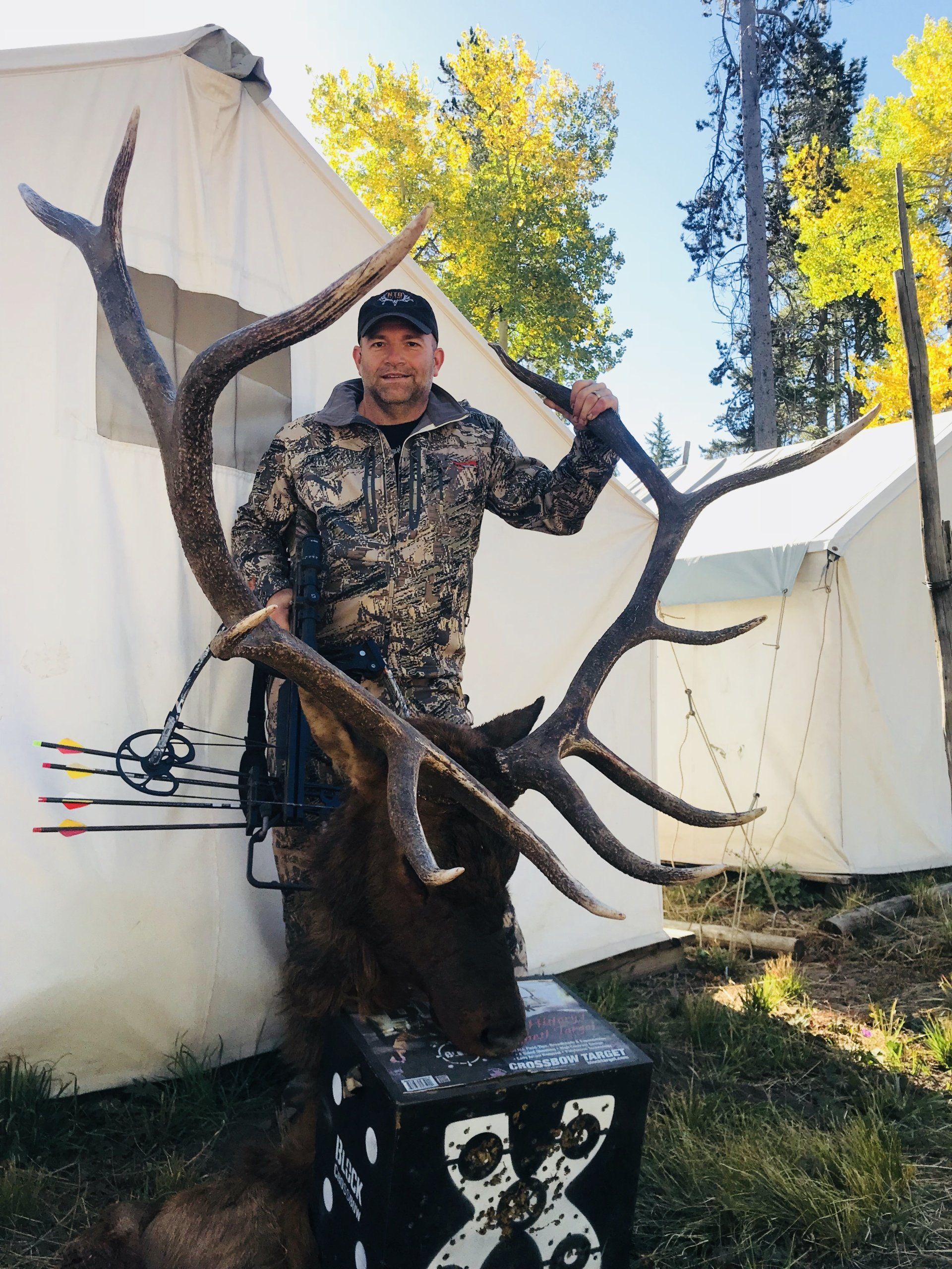 Man in camo holds large elk antlers, posing by a tent in a wooded area.