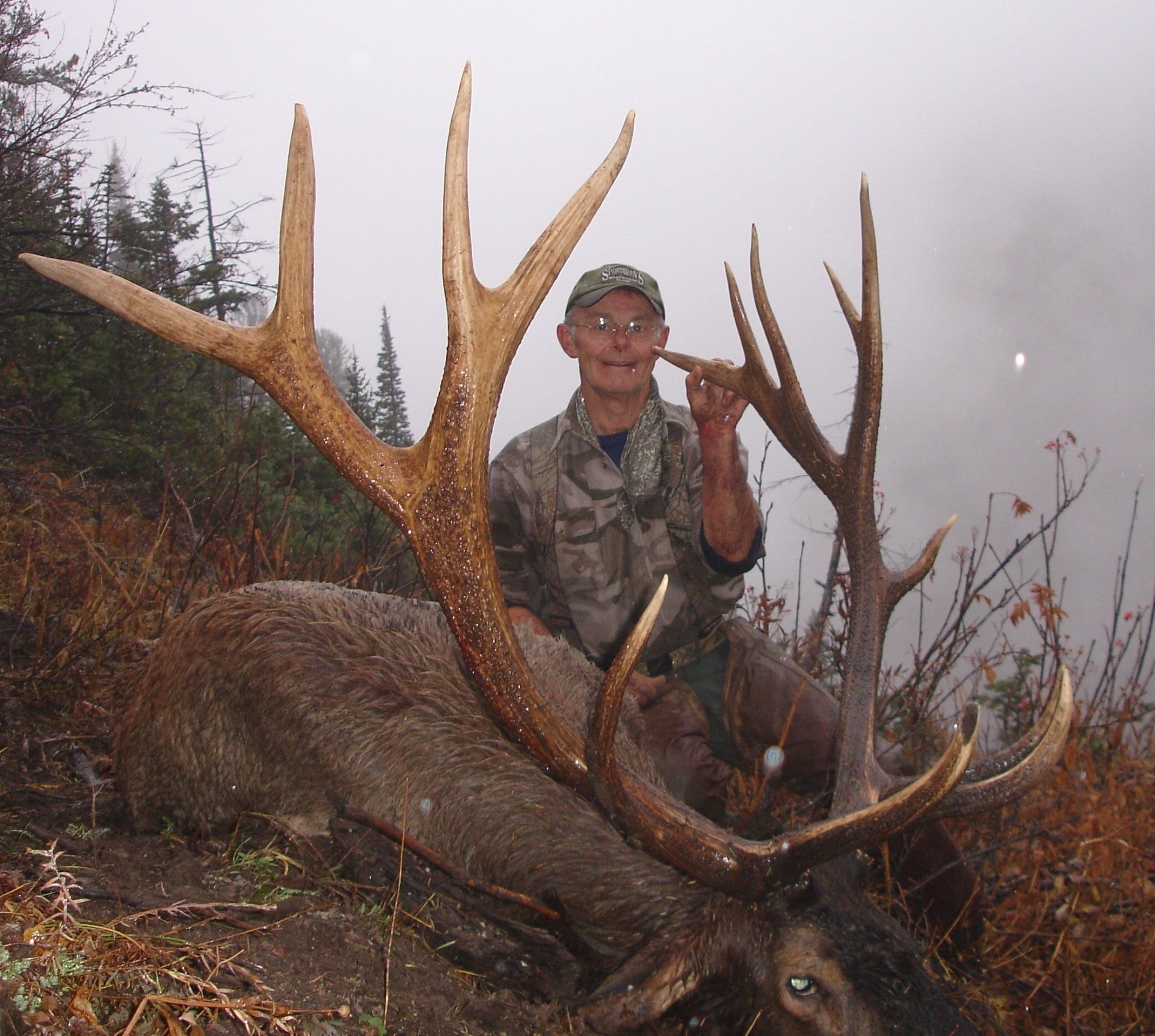 Hunter smiles next to a large elk with impressive antlers in a wooded area.