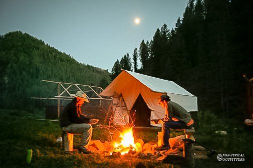 Two men sit around a campfire in front of a tent