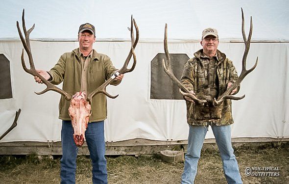 Two men hold up large elk antlers and a skull in front of a white building.