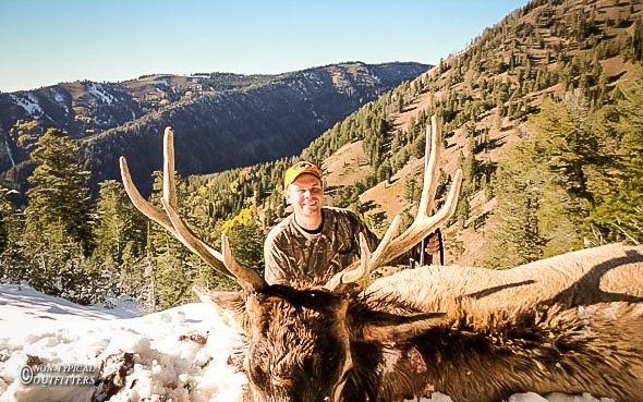 Man in camouflage with elk, mountains in the background. Snow on ground.