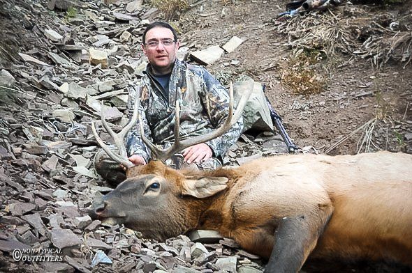Man in camo with elk he shot in rocky terrain. Elk has large antlers.