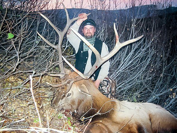 Hunter kneels beside a large elk with antlers. He has one hand raised above head in the woods.