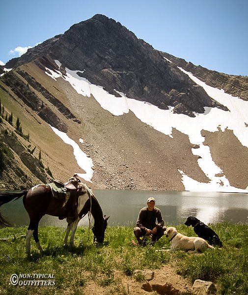 Man with two dogs and horse by a lake, mountain backdrop with snow.
