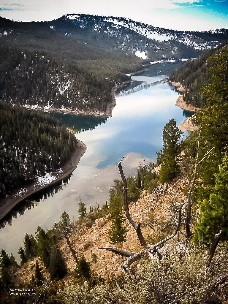 A scenic view of a lake in a mountain valley, with snow-covered banks and forested slopes.