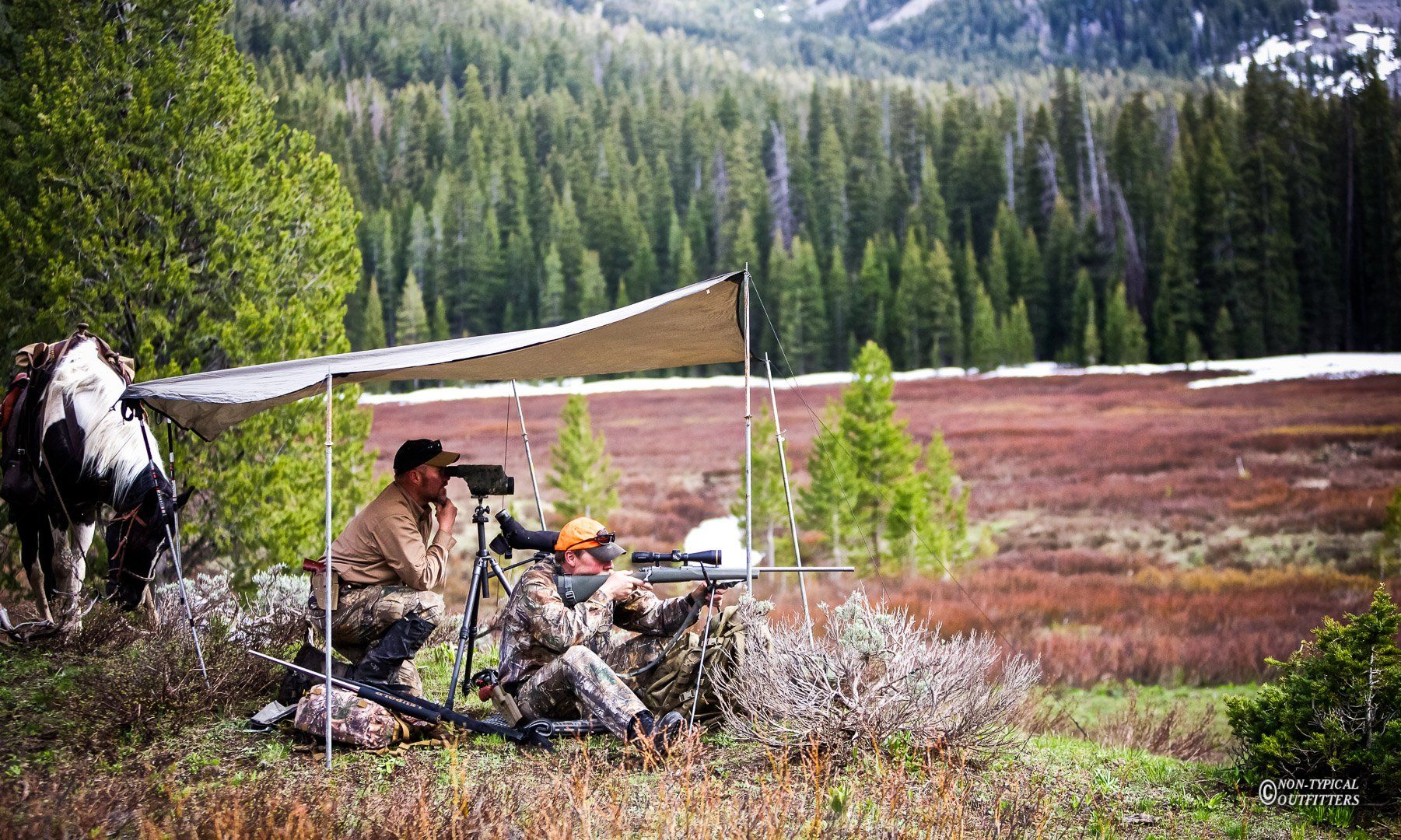Two men are sitting under a tent looking through binoculars