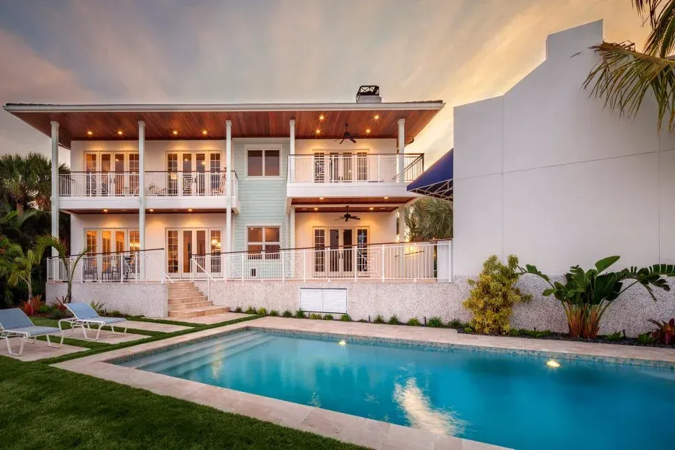 Two-story white house with balconies and pool at dusk; blue pool, lounge chairs, and greenery.