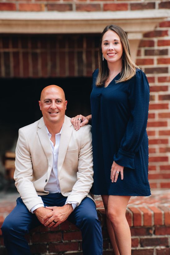 Man seated, woman standing; pose near brick fireplace. Man wears tan jacket, blue pants; woman in navy dress.