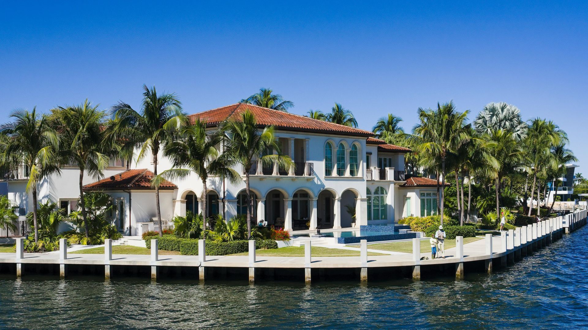 White mansion on a canal, palm trees, blue water, sunny day.